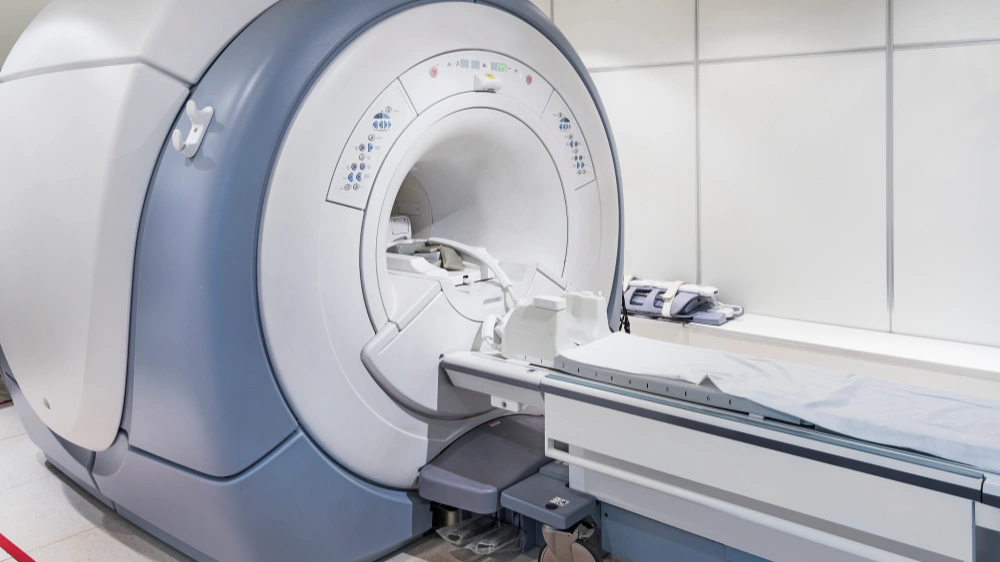 A photo of a bright and clean hospital examination room. The main subject is a large MRI (Magnetic Resonance Imaging) scanner, featuring a white and light blue-gray exterior with a characteristic large circular bore. Inside the opening, a dedicated head coil is installed for brain imaging. A patient bed with a light blue sheet extends in front of the machine for patients to lie on. On the white counter to the right, a folded gray blanket or patient gown is neatly placed. The room is well-lit, giving off a sterile and modern medical atmosphere.
