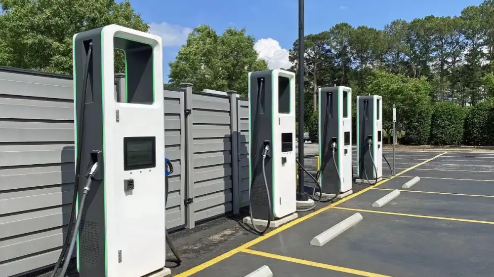 An outdoor parking lot photo showing four identical white electric vehicle charging stations lined up side by side. The chargers feature black front panels, screens, and green trim. They are installed in front of a gray metal fence, with yellow parking lines and white concrete wheel stops on the ground. The background shows dense green trees and a clear blue sky with a white cloud.