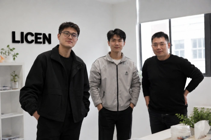 A photo of three Licen team members posing together in the office. The man on the left wears glasses and a black jacket, the middle man wears a light grey color-block jacket, and the man on the right wears a black crewneck sweater. They are standing and smiling in front of a white wall displaying the 'LICEN' logo, with a plant shelf and a window nearby.