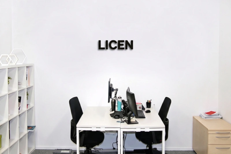 A bright, minimalist office space featuring a white wall with black 'LICEN' lettering, two desks with computers and black chairs, a white shelving unit, and a small wooden filing cabinet.