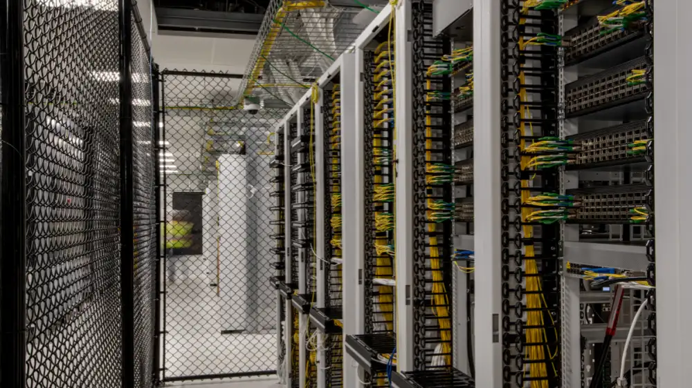 An indoor photo of a data center showing a row of white server racks lined up closely together. The racks are packed with networking equipment, and the vertical cable organizers are filled with neatly bundled yellow and green network cables. A black wire mesh security fence runs along the left side and front of the aisle. The background shows bright, cool-toned overhead lighting, creating a clean, orderly, and high-tech atmosphere.