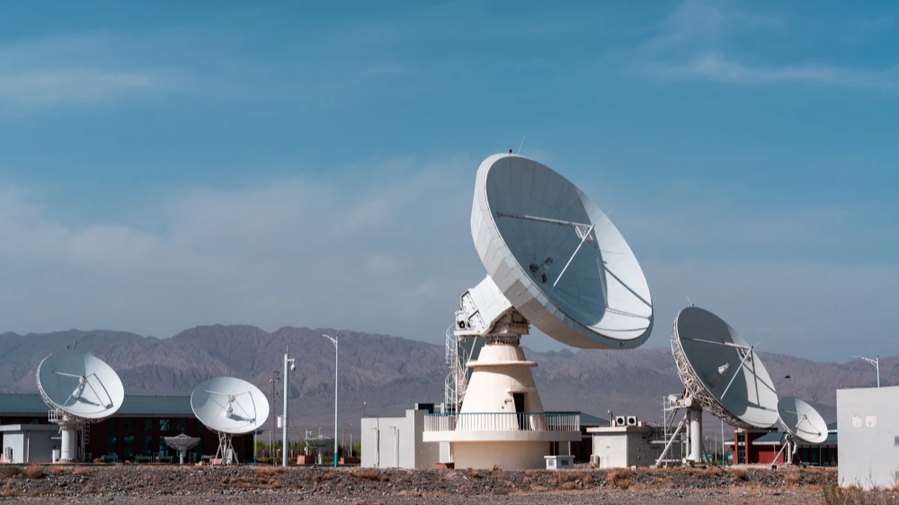 Multiple white parabolic antennas form a satellite ground station on an arid gravel plain. In the center is a large dish mounted on a cylindrical pedestal with railings, surrounded by several smaller antennas. The background features rugged brown mountains and a clear blue sky, with small buildings and light poles visible on site — a typical remote communication facility.