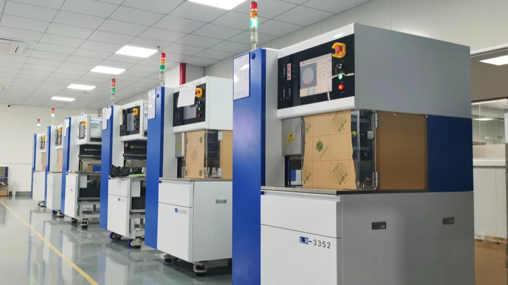 A row of blue and white wafer dicing machines in a cleanroom production line, each with a status light tower on top, a touchscreen control panel, and an emergency stop button. Some machines have cardboard covering the middle sections. The model number 'LX-3352' is visible on the base. The floor is glossy with a yellow safety line.
