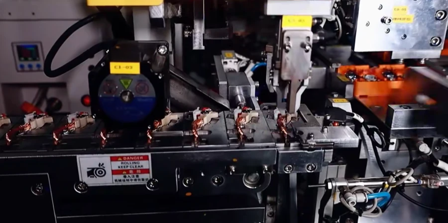A close-up of an automated production line assembling miniature circuit breakers, with copper coil components held in a metal track, and a DANGER rolling keep clear warning label on the machine front.