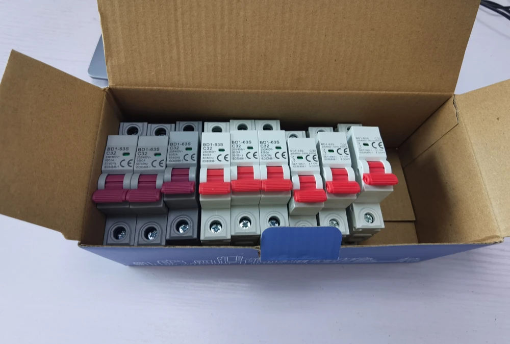 An open brown cardboard box filled with neatly arranged MCBs. The three units on the left feature a grey body and pinkish-red handle, while the units on the right have a white body and red handle. The front of the breakers clearly shows the 'BD1-63S C32' model number and CE certification mark.