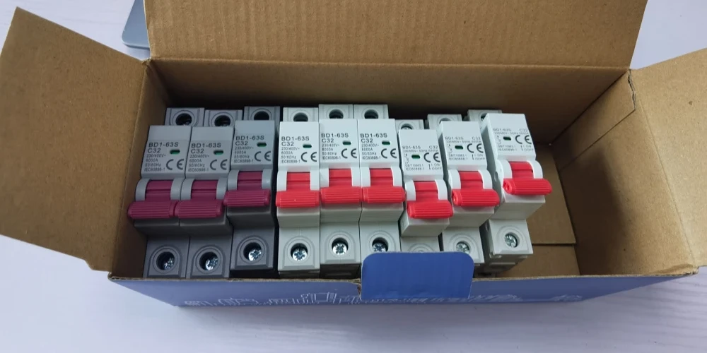 Top-down view of an open cardboard box containing 9 neatly arranged miniature circuit breakers in grey and white casings. The switches feature pink and red levers, printed with 'BD1-63S C32' model specifications and CE certification marks, with a blue label on the front edge of the box.