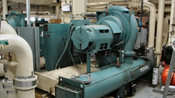Core chiller unit for commercial HVAC systems, teal main unit mounted on a concrete base in a mechanical room, connected to large insulated water pipes and electrical control equipment