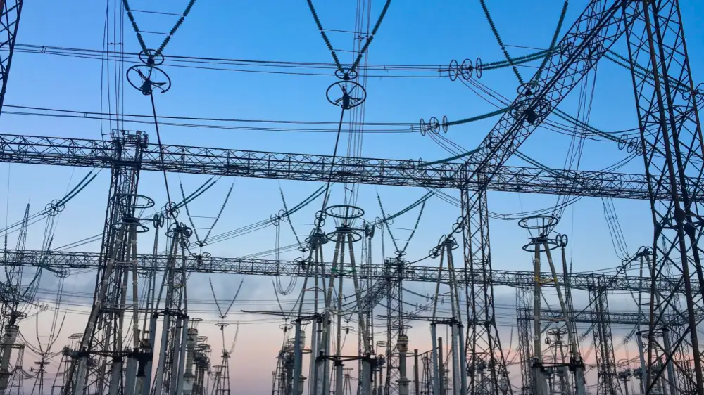 A low-angle image showing the intricate network of transmission lines, insulators, and towering steel frameworks within a DC distribution system. Set against a gradient sky at sunrise or sunset, the photo highlights the dense electrical connections and infrastructure inside a DC substation or converter station.