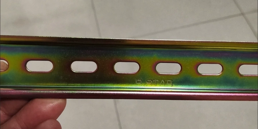 A close-up photo of a hand holding a color zinc-plated DIN rail. The metal surface shows an iridescent yellow-green finish, with a row of elongated mounting holes evenly spaced along the middle. This is a standard mounting track commonly used in electrical panels to secure miniature circuit breakers.