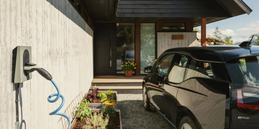 A wall-mounted electric vehicle charger with a coiled blue cable on a concrete wall, next to a raised planter bed and a black car parked in a driveway in front of a modern house.