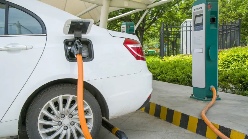 A close-up image showing a white electric vehicle parked under an outdoor canopy, with a thick orange charging cable connected to its charging port. The car is plugged into a green-and-white DC fast charging pile nearby. The surrounding environment includes green shrubs and a metal fence.