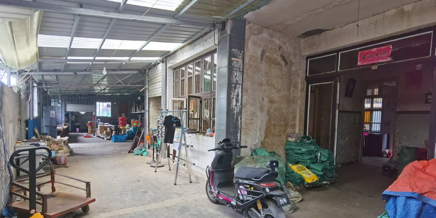 Ground-floor converted workshop with a metal roof, a parked scooter, stacked green sacks, and storage materials, typical of a small family business.
