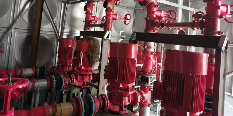 A row of red fire pumps connected to red pipes and valves, installed in a room with large silver water tanks in the background.