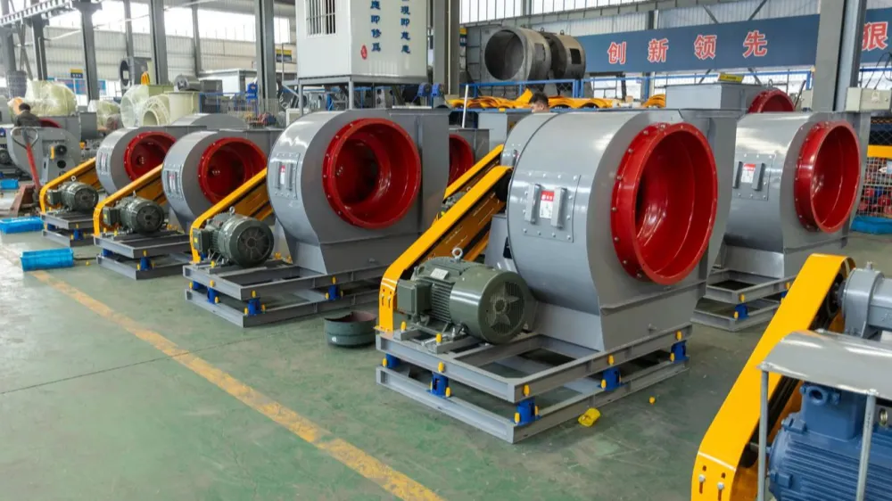 Row of gray industrial centrifugal fans with red inlets, green motors, and yellow drive belts in a factory workshop