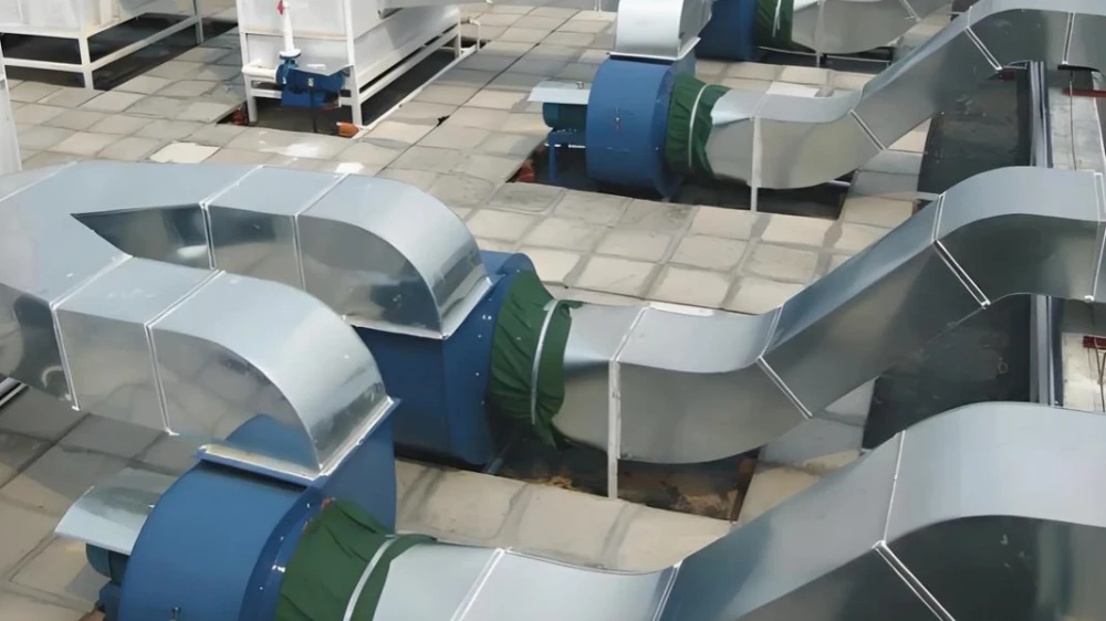 Rooftop industrial ventilation and exhaust system, with multiple blue centrifugal fans connected to silver square galvanized ducts via green flexible connectors, well-organized layout.