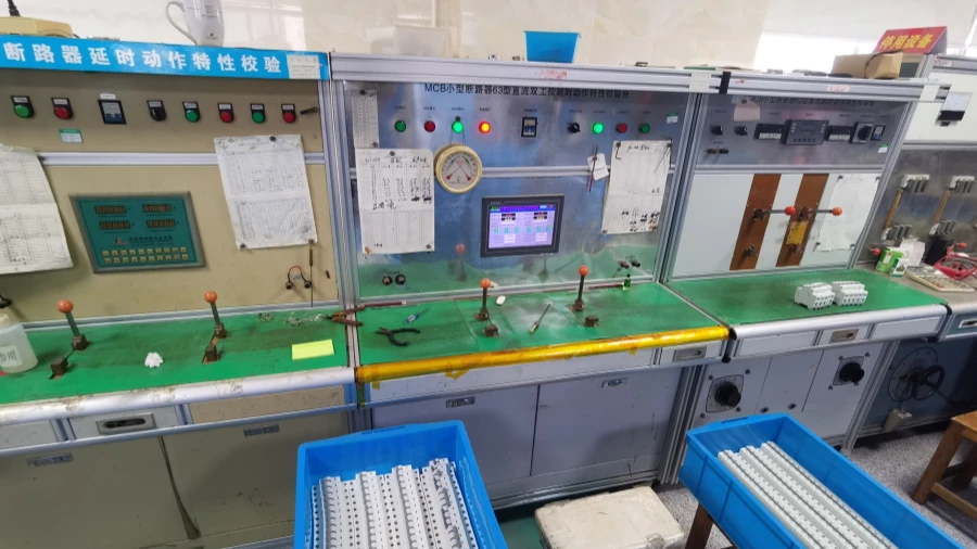 Industrial verification workstations for miniature circuit breakers with green countertops, control panels, switches, digital displays, and blue bins holding white MCBs in the foreground