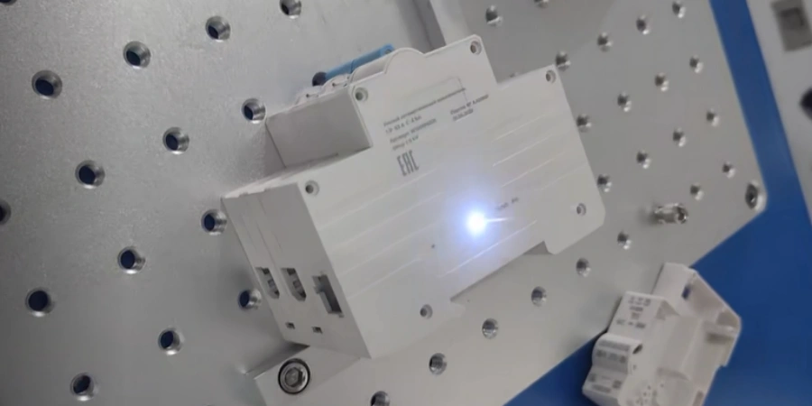 On a metal laser marking workbench covered with positioning holes, a white miniature circuit breaker plastic housing is placed. The surface has been clearly marked with the brand logo and product parameters via laser marking. A bright white laser spot generated during the marking process illuminates the surface. Another white breaker housing waiting to be marked is partially visible in the bottom right corner.