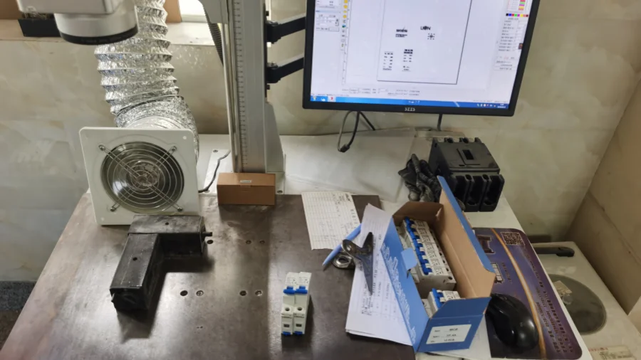 A workbench with a computer monitor, laser marking equipment with exhaust ducting, a test fixture, papers, a mouse, and an open box containing miniature circuit breakers