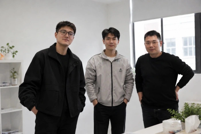A photo of three Licen team members posing together in the office. The man on the left wears glasses and a black jacket, the middle man wears a light grey color-block jacket, and the man on the right wears a black crewneck sweater. They are standing and smiling in front of a white wall displaying the 'LICEN' logo, with a plant shelf and a window nearby.