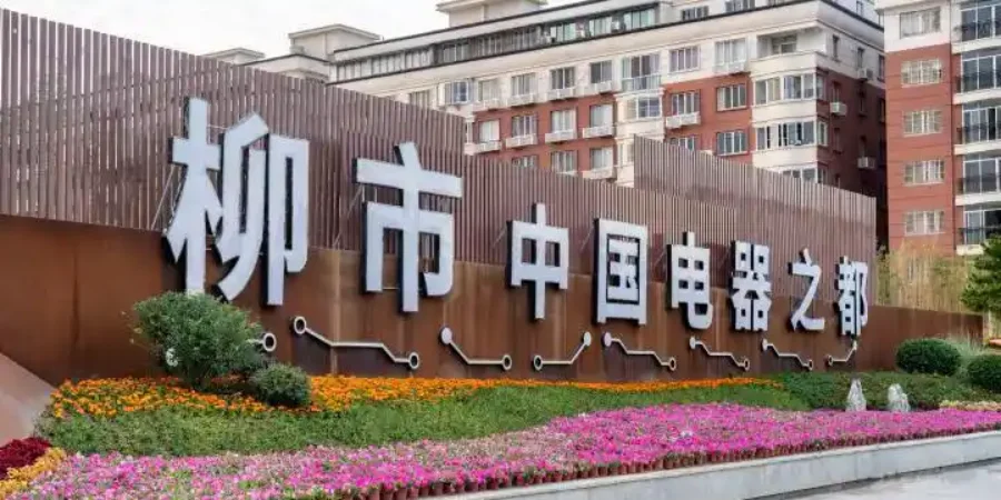 Large white Chinese characters on a brown metal structure reading Liushi China Electrical Appliance Capital, with a flower bed in front and residential buildings behind.