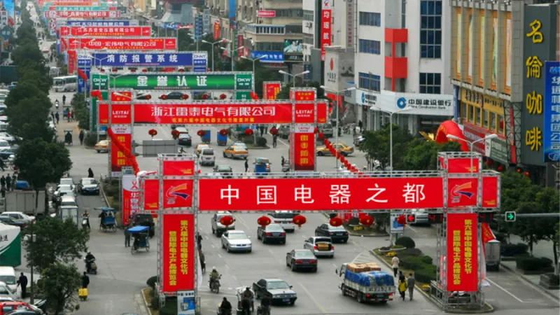 A busy street in Liushi town with heavy traffic, lined with large red banners and arches featuring Chinese text, alongside buildings and pedestrians.