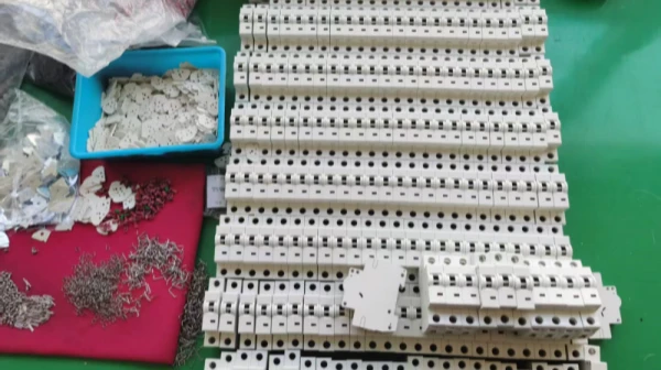 A row of white plastic MCB housings lined up on an assembly table. On the left, there is a blue bin with plastic components, and a red mat with scattered metal internal parts for the circuit breakers.