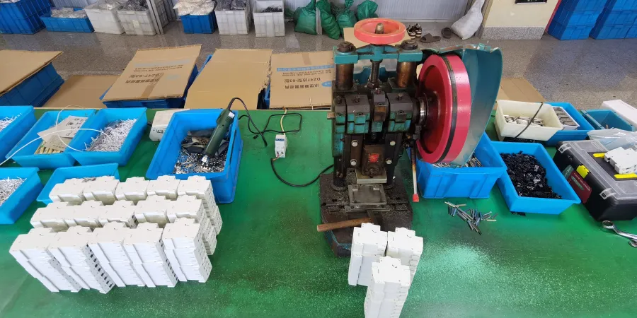 A manual MCB riveting press on a green workbench, surrounded by blue bins of metal parts and stacks of white circuit breaker housings.