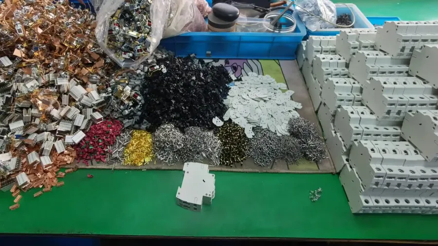 A green workbench with piles of metal parts, screws, plastic pieces, and stacked white MCB casings
