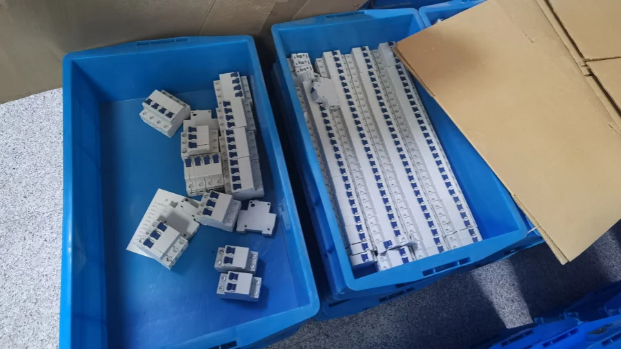 Two blue plastic bins on a floor, one holding loose white miniature circuit breakers and the other containing rows of connected white MCBs, with cardboard beside them