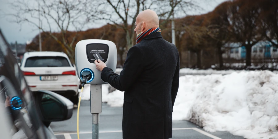 A bald man in a black coat and striped scarf stands at a white pedestal EV charger, holding a card over the reader, with a plugged-in cable and snowbanks in the background.