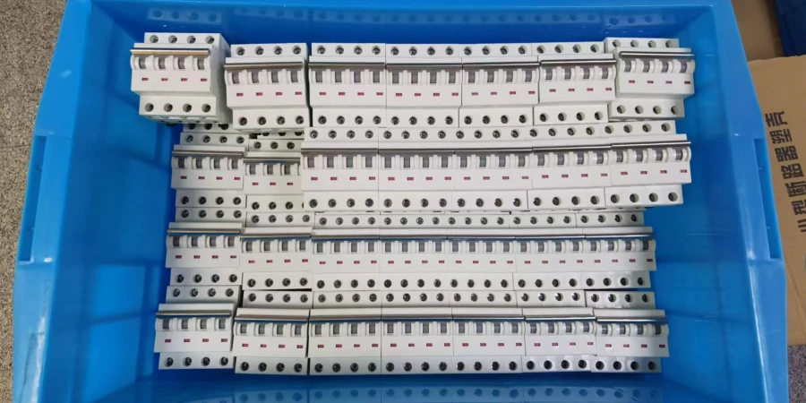 An overhead photograph showing a blue plastic storage bin filled with neatly arranged rows of white MCBs. The breakers are predominantly configured as three-pole units, featuring small red indicators on their side panels and visible black terminal screw holes on the front. On the right edge of the frame, a portion of a cardboard box is visible, displaying black Chinese characters.