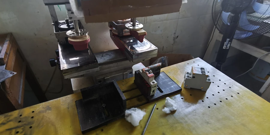 On a yellow workshop workbench, a pad printing machine with red silicone pads is set up for operation. The table holds mcbs ready for pad printing, a black base, and a gray semi-finished component. White debris and a metal tool are scattered nearby for adjustment, with an industrial fan visible in the background on the right.