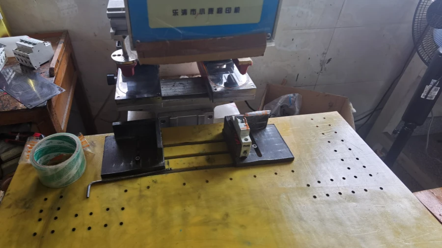 A pad printing machine on a yellow workbench with a miniature circuit breaker positioned on a black fixture, with tape and other tools nearby