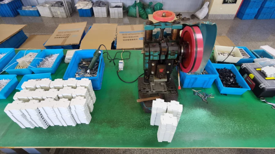 A green workbench with a riveting press machine in the center, stacks of white MCB casings beside it, and blue bins holding small metal parts and tools around the table