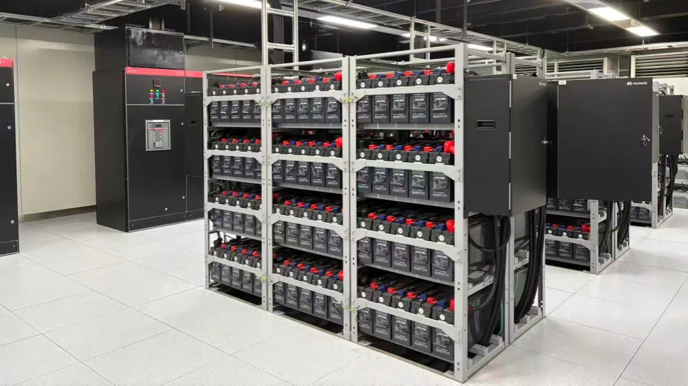 UPS uninterruptible power supply system installed in a data center server room, including multi-tier battery banks on metal racks and a large black distribution cabinet on the left, with neatly routed cables