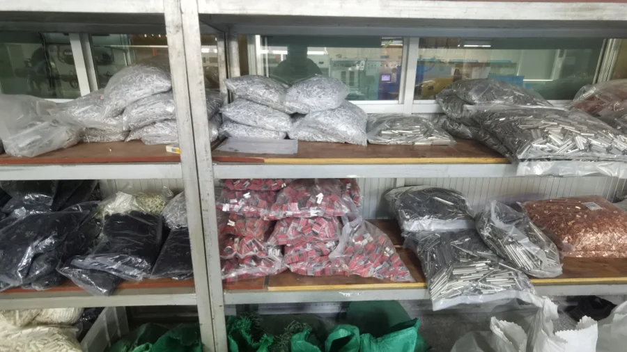Metal shelving units holding clear plastic bags filled with various miniature circuit breaker components, including silver, black, red, and copper-colored metal parts.