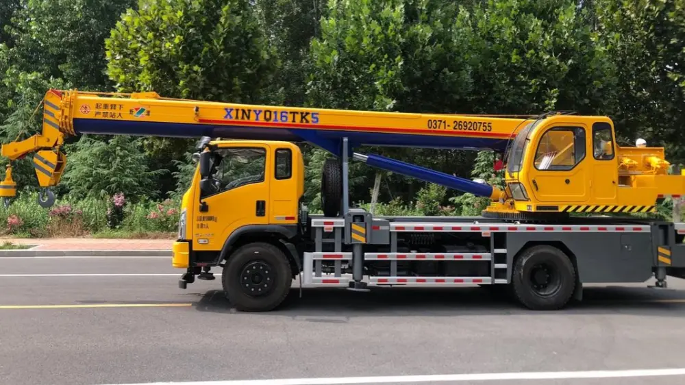 Yellow and blue truck crane, side view showing telescopic boom, dual cabs, and safety warning markings on the body, parked on factory road