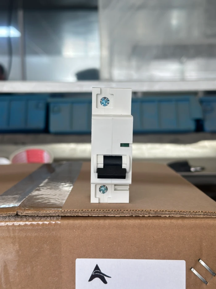 A white MCB standing vertically on top of a brown corrugated cardboard box. The breaker has a black toggle handle and metal screw terminals at the top and bottom. The background features blurred blue storage cabinets. On the surface of the box, there is a strip of transparent tape and a white label printed with a black triangular logo.