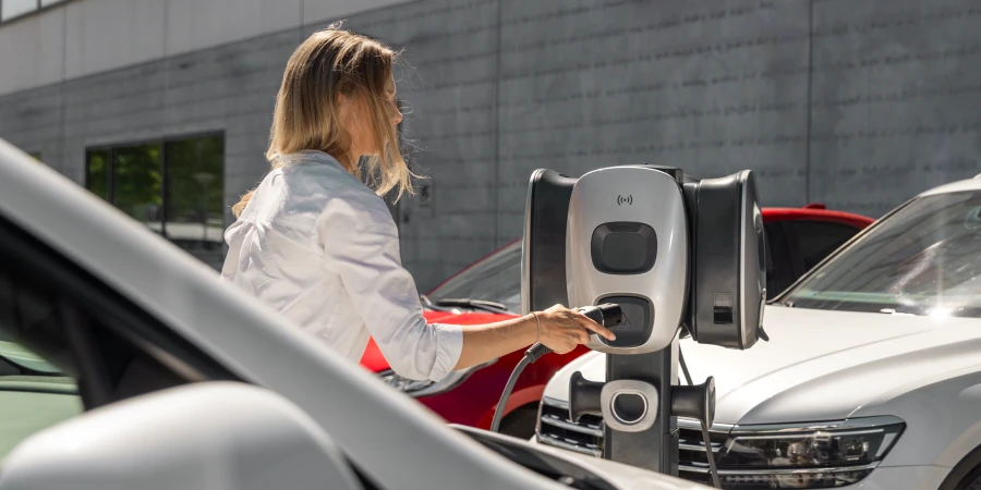 A woman in a white shirt stands at an outdoor charging station and inserts a charging plug into a silver and black pedestal charger, with parked cars in the background.