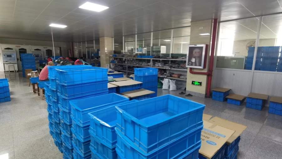 A bright workshop floor with large stacks of blue plastic bins in the foreground, workstations and workers in the background, and a fire extinguisher cabinet on the wall