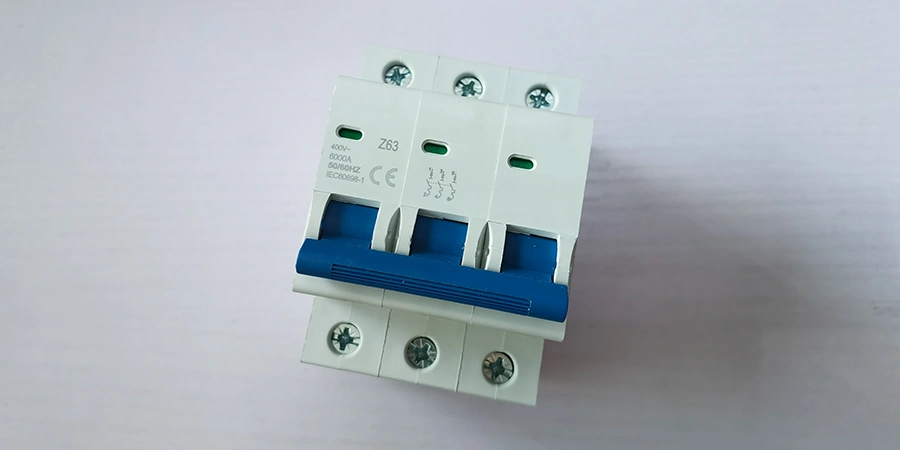 A white three-pole Z curve miniature circuit breaker with a blue handle and green indicator windows, placed on a white desk.