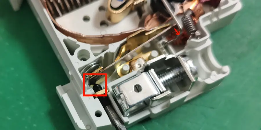 A close-up of the internal mechanism of an mcb on a green workbench, showing a brass bimetallic strip, copper wire, a small calibration screw highlighted in a red box, a red arrow pointing to a spring, and a threaded adjustment screw.