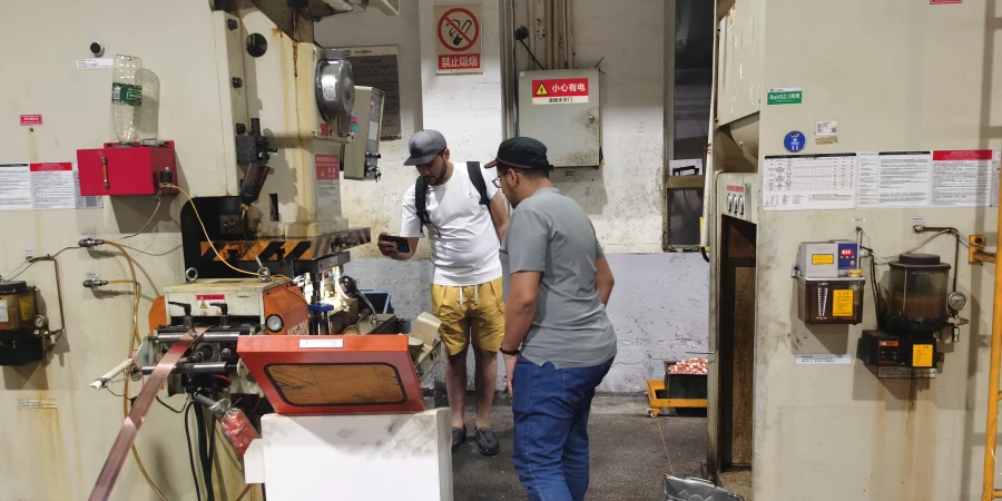 Two Algerian clients in casual clothes stand in a factory workshop, one holding a phone, looking at an industrial press machine with warning signs and control panels on the walls.