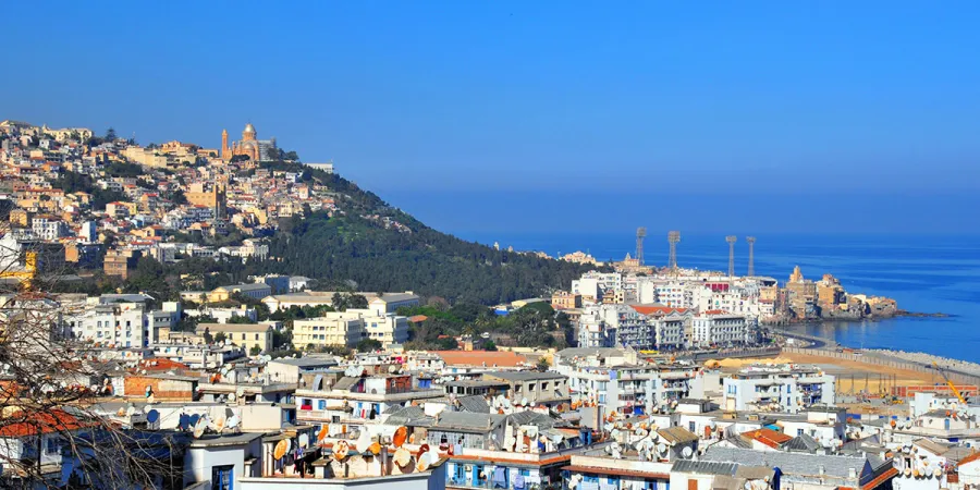 A view of Algiers with white buildings spread across a hillside and along the coast, a large domed structure on the hill, and the blue sea in the background