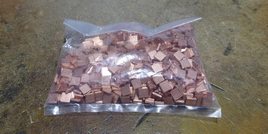 A clear plastic bag filled with small copper-colored contact pieces, many with a textured grid pattern on top, resting on a worn metal surface