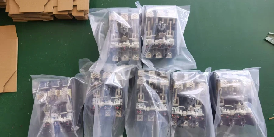 Several clear plastic bags filled with MCCB semi-knocked-down components, placed on a green workbench next to stacked cardboard boxes.