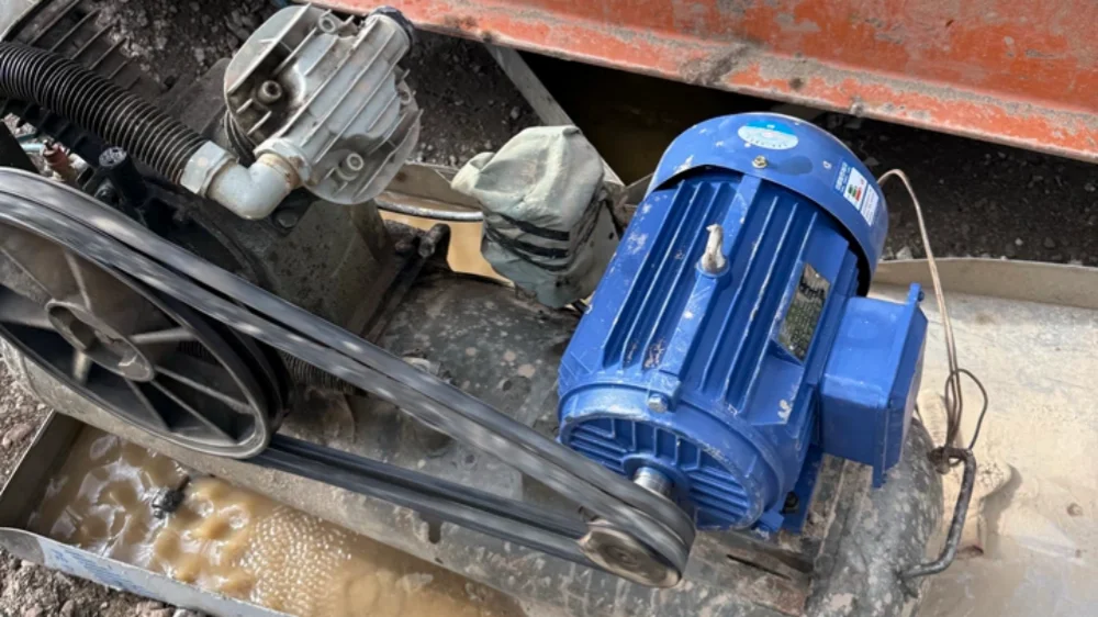 A blue electric motor connected by belts to a large pulley wheel, mounted on a dirty metal frame with mud and debris underneath.