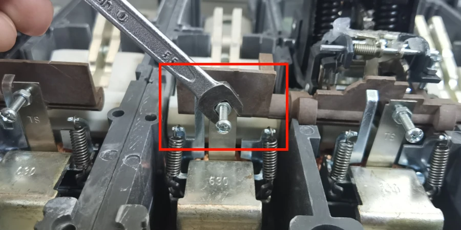 A hand holds a metal wrench next to a fixed calibration screw highlighted in a red box on a molded case circuit breaker, with metal brackets, springs, and stamped numbers visible in the background.
