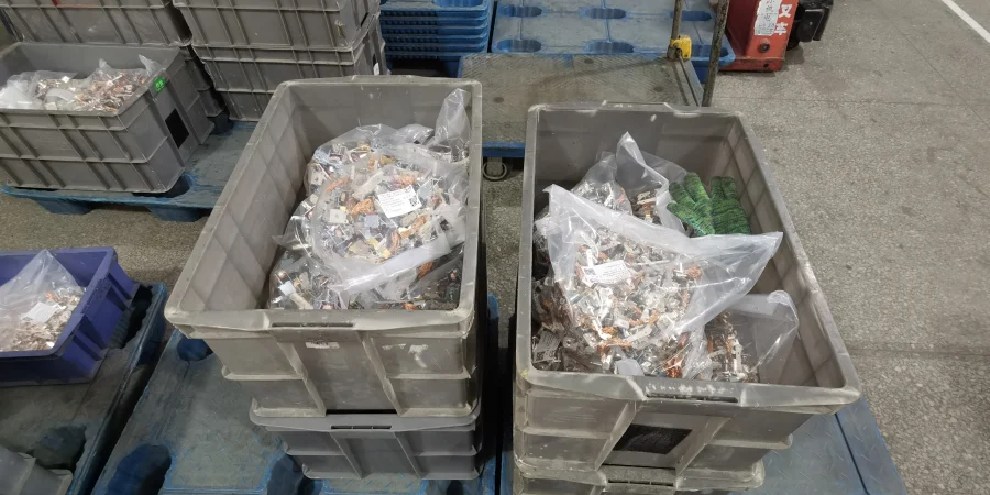 Two large gray plastic bins sit on a warehouse floor, each holding clear plastic bags filled with small circuit breaker components. More gray and blue bins are stacked in the background.