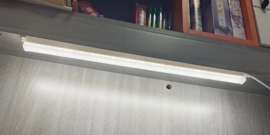 A white LED light bar mounted under a gray shelf, illuminating a wall with a herringbone pattern. A small round hole is visible on the wall below the light. Books and boxes sit on the shelf above.